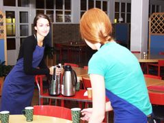 Två unga kvinnor i förkläden hanterar kaffetermosar och koppar i en kafeteria.