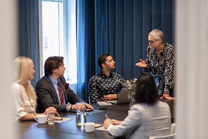 En grupp människor har ett möte runt ett konferensbord. En kvinna står och pratar medan de andra sitter och lyssnar.