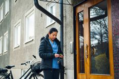 En kvinna som arbetar inom hemtjänsten står utanför en port till ett flerfamiljshus. Hon håller i en nyckelklippa. Bakom henne står en cykel.