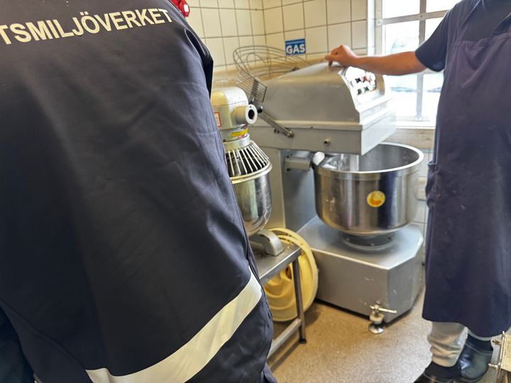 Bild på en arbetsmiljöinspektörs rygg och en degblandare i bakgrunden tillsammans med en arm tillhörande en arbetstagare.