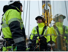 Två personer med gula skyddsjackor och personlig fallskyddsutrustning arbetar på höjd vid en byggnad.