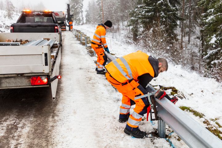 Arbetare i orange varselkläder reparerar ett vägräcke vid en snötäckt väg.