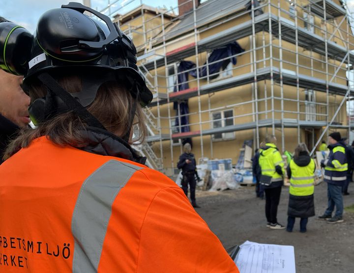 Arbetsmiljöinspektörer står med ryggen empt fotografen i för- och bakgrund på en byggarbetsplats med ställningar på en husvägg.