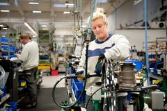 En kvinna med skyddsglasögon och skyddshandskar arbetar med en maskin och en man i bakgrunden arbetar vid en arbetsbänk.