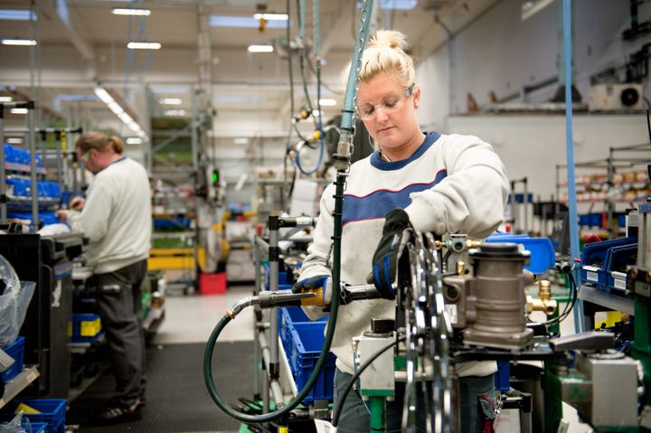 En kvinna med skyddsglasögon och skyddshandskar arbetar med en maskin och en man i bakgrunden arbetar vid en arbetsbänk.