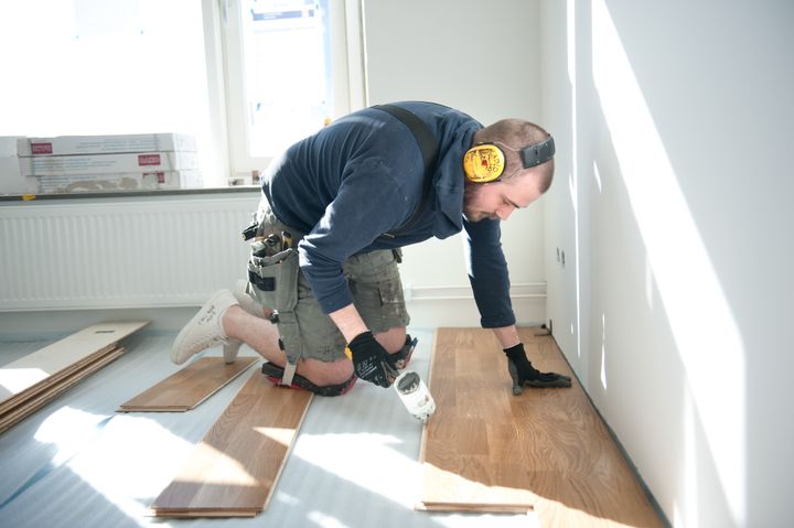 En person installerar trägolv inomhus, och bär hörselskydd och en verktygsbälte.