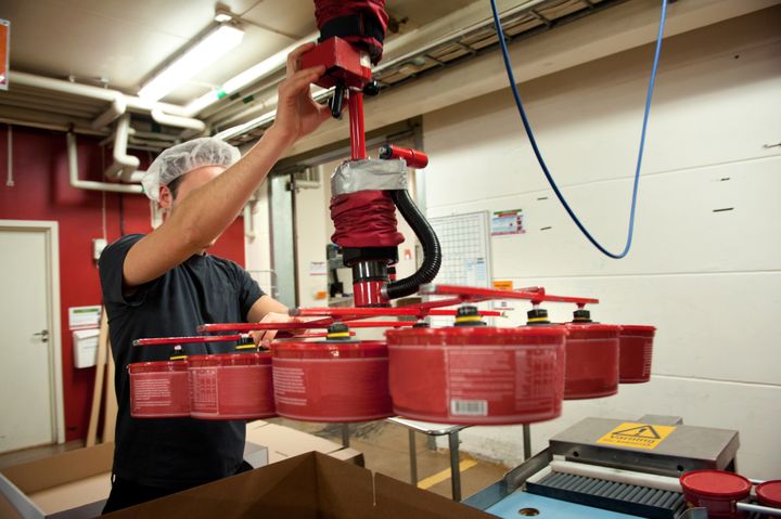 En person med hårnät använder en maskin för att lyfta flera röda burkar på en fabrik.