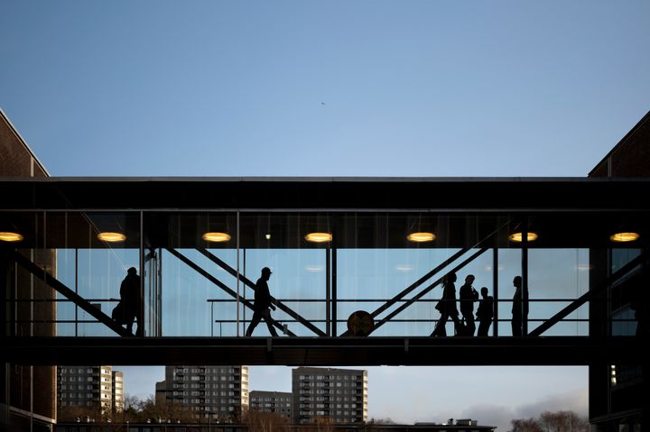 Personer går över en inglasad gångbro mot en klarblå himmel, med stadssilhuett i bakgrunden.