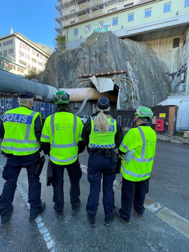 Alt text: Fyra personer i uniform står framför en byggarbetsplats med en tunnelinngång vid bygget av Västralänken i Göteborg.