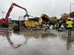 En skrotningsplats med en grävmaskin med gripklo som lyfter metallskrot. Containrar och metallavfall ligger utspridda på en våt gårdsplan. Flera personer i varselkläder står i förgrunden.