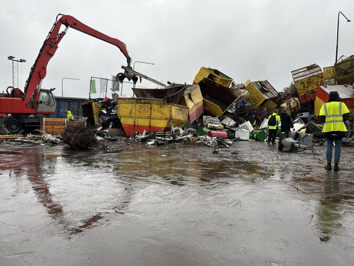 En skrotningsplats med en grävmaskin med gripklo som lyfter metallskrot. Containrar och metallavfall ligger utspridda på en våt gårdsplan. Flera personer i varselkläder står i förgrunden.