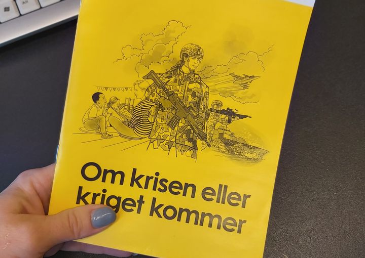 Framsidan av den gula foldern med rubriken "Om krisen eller kriget kommer" hålls av en hand mot bakgrund av grå skrivbordsyta och en skymt av ett tangentbord.