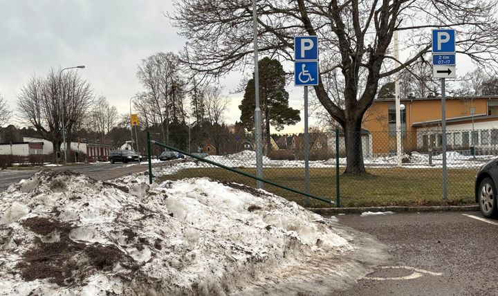 Snövall framför blåvit skyltning som markerar p-plats för trafikanter med funktionsnedsättning. Till höger syns att den allmänna parkeringen är snöfri. I bakgrunden träd och byggnader, i stort sett barmark.