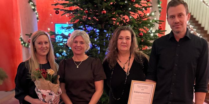 Fyra glada personer står framför en julgran och håller i blommor och ett diplom.