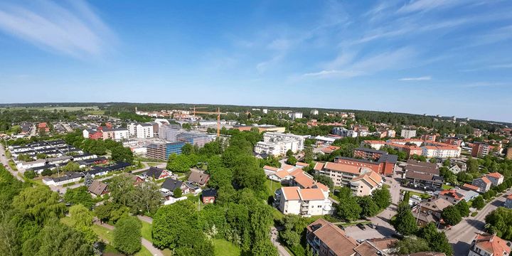 Flygfoto av en stad med bostadsområden, grönområden och skog i bakgrunden under en klarblå himmel.