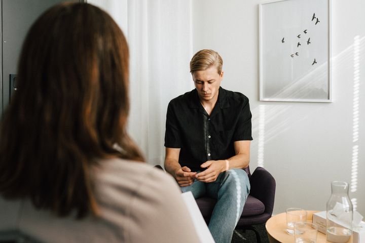 Ung man i samtal med kvinna som bara syns bakifrån. Mannen ser ner på sina händer. Miljön liknar rum för samtalsterapi.