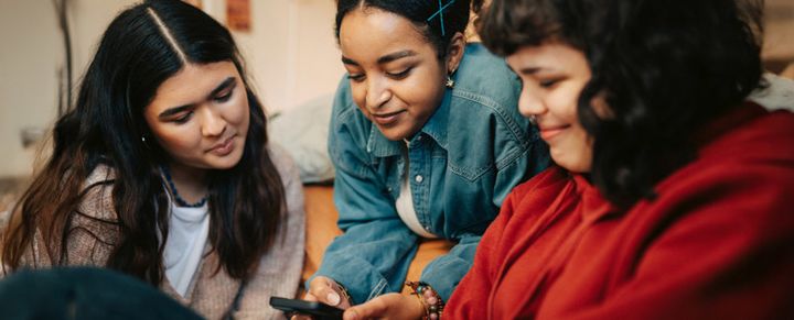 Tre unga kvinnor sitter tillsammans och tittar engagerat på en mobiltelefon.