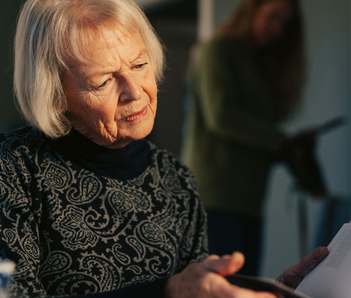En äldre kvinna sitter och tittar i en bok. I bakgrunden syns en yngre kvinna sträcka ner en hand i en väska samtidigt som hon tittar mot den äldre kvinnan.