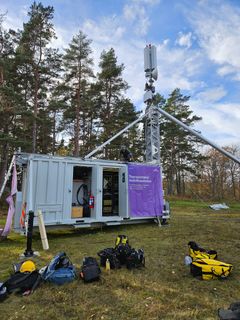 Mobil telemast på fält med utrullad teknik från container, omgiven av träd och utrustning på marken.