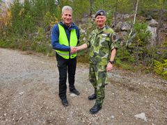 Två män skakar hand utomhus. En har en gul väst, och den andra är klädd i militäruniform.