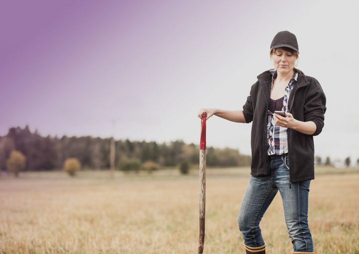 Kvinna på en åker med en spade i ena handen och mobiltelefon i den andra.