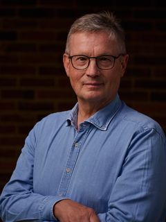 Man i blå skjorta och glasögon står framför en mörk bakgrund.