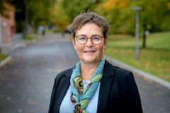 Porträttfoto av Annika Olsson, kvinna med kort brunt hår och glasögon, klädd i mörk kavaj och mönstrad sjal, stående utomhus med en parkväg och höstlöv i bakgrunden.