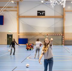 En flicka i en idrottshall kastar en basketboll mot en basketkorg medan barn leker i bakgrunden.