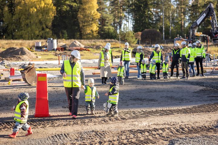 Förskolebarn och pedagoger med byggvästar och hjälmar går tillsammans på en byggarbetsplats.