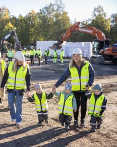 Förskolebarn och pedagoger med hjälmar och västar promenerar över en byggarbetsplats.