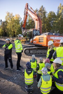 Förskolebarn och pedagoger med byggvästar och hjälmar tittar på en grävmaskin vid en byggarbetsplats.