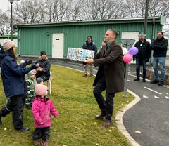 En man ger ett paket till en kvinna utomhus, omgiven av barn och vuxna. Ballonger hänger på en stolpe.