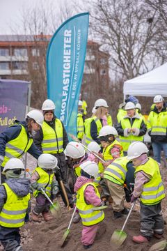 En grupp barn i reflexvästar och hjälmar gräver med spadar i sanden. Vuxna i liknande kläder står runt omkring.