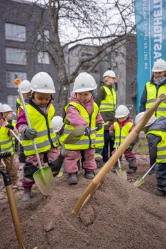 Barn med hjälmar och reflexvästar leker i en sandhög med spadar på en byggarbetsplats.
