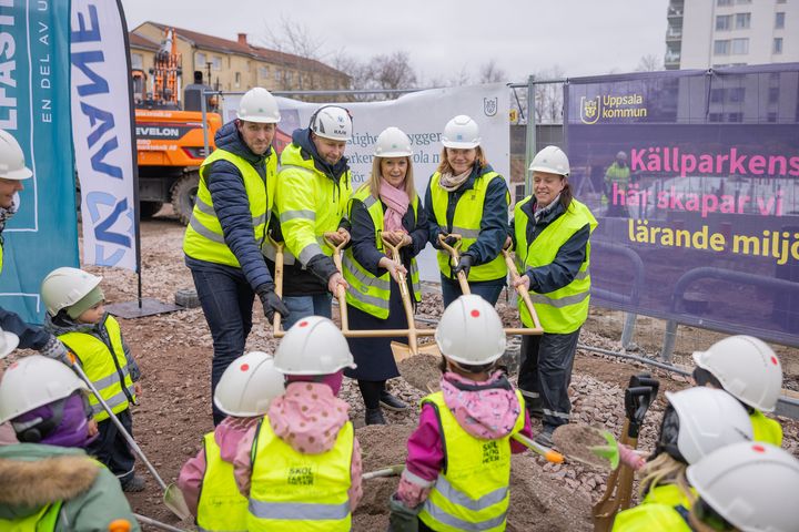 En grupp vuxna i skyddshjälmar och västar deltar i en spadtagsceremoni tillsammans med barn i liknande utrustning på en byggarbetsplats.