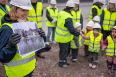 Person i bygghjälm och reflexväst håller upp en bild, omgiven av flera vuxna och barn som också bär hjälmar och reflexvästar.