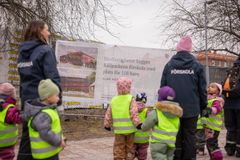 Grupp av barn och vuxna i reflexvästar promenerar utomhus vid en byggarbetsplats med en skylt som annonserar ny förskola.