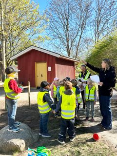 En grupp barn i reflexvästar lyssnar på två vuxna som pratar om träd på en skolgård.