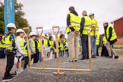 En grupp människor, inklusive barn, i byggkläder med hjälmar och reflexvästar, står framför en rad spadar på en byggarbetsplats.