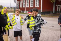 Tre barn provar reflexvästar utomhus nära en röd byggnad.