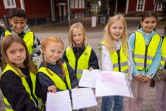 En grupp barn i reflexvästar håller papper utomhus.