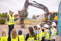 En man i reflexväst och hjälm pratar med en grupp barn i reflexvästar framför en grävmaskin.