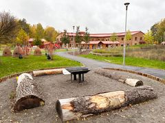 Lekplats med stockar och grillplats framför en tegelbyggnad i höstmiljö.