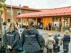 Barn och förskolepersonal samlade på en förskolegård framför en träbyggnad.