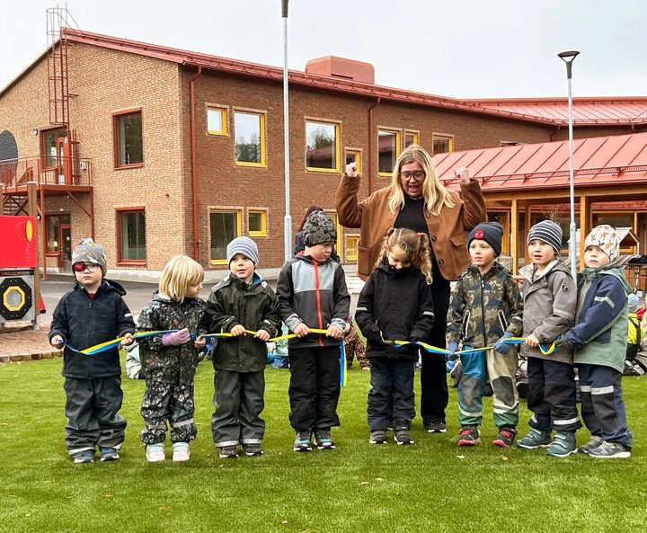 En grupp förskolebarn klipper ett blågult band framför en skolbyggnad, tillsammans med en vuxen.