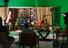 En man sitter på en stol i en inspelningsstudio med bakgrund av en bokhylla och kamerautrustning.