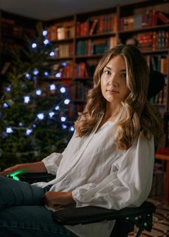 En person med långt hår sitter i en stol framför en bokhylla och en julgran med ljus.