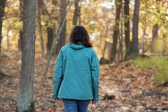 En person i mörkt, långt hår iklädd höstjacka, promenerar genom skogen, med ryggen mot kameran.