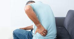 En man med ont i ryggen sitter på en soffa och tar sig för ryggen där det gör ont.