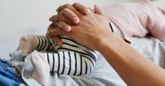 En förälder med sin nyfödda bebis ligger på rygg med bebisen ovanpå magen. Föräldern håller med knäppta händer om bebisen som ligger på förälderns mage. Bebisen har randiga byxor och en rosa tröja på sig.